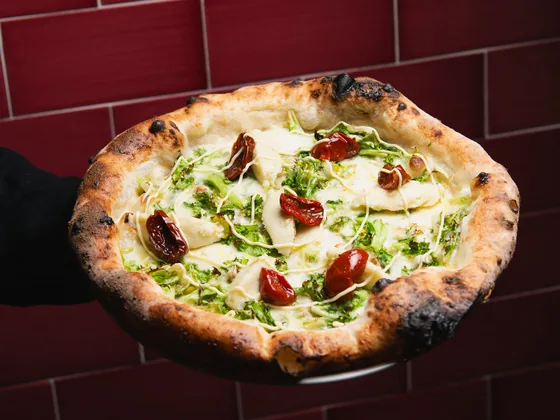 Una pizza bianca con datterini confit tenuta sul palmo davanti alle piastrelle bordeaux