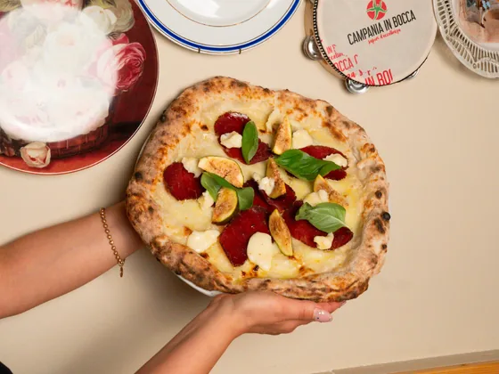Una pizza portata a due mani sopra il tavolo, accanto ai piatti di ceramica del locale